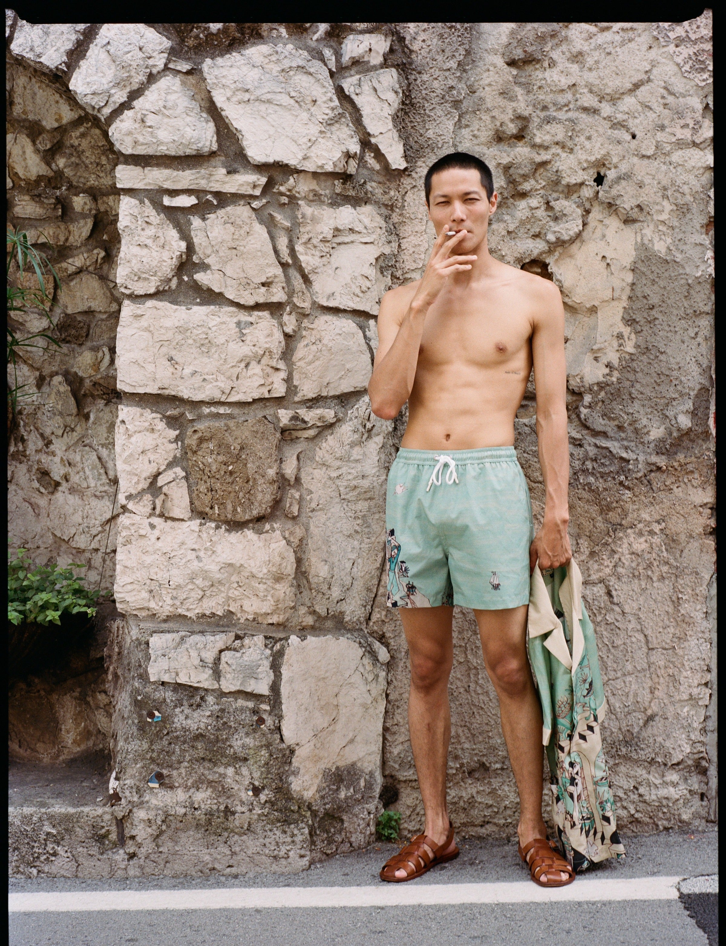 Male model wearing swim trunks and sandals smoking a cigarette