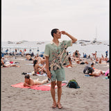 Man wearing matching Liguori shirt and swim trunks holds hand over his eyes and gazes off into the distance amidst a crowded beach