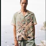 Man stares at camera with Amalfi bay in the background wearing the silk Liguori shirt and matching swim trunks