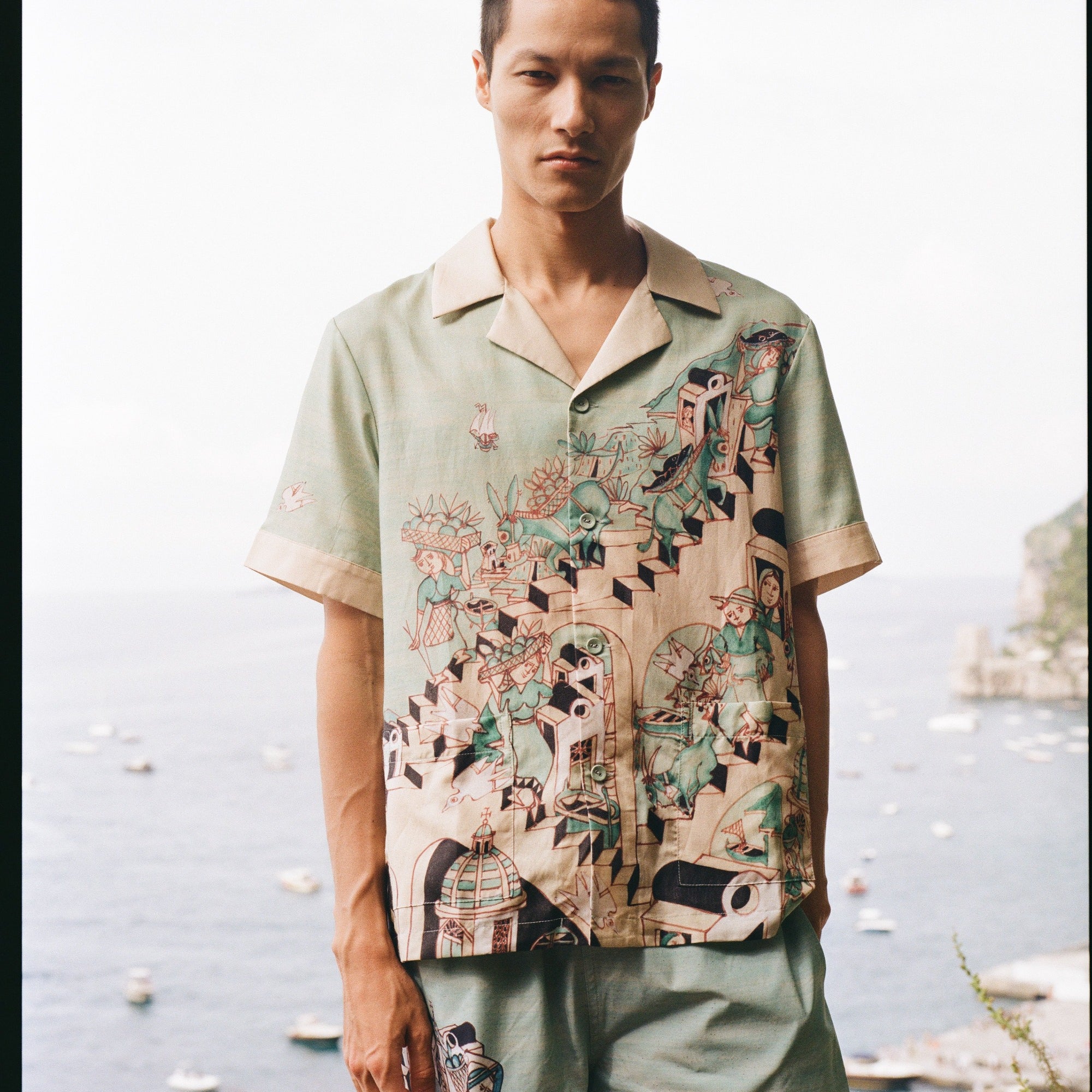 Man stares at camera with Amalfi bay in the background wearing the silk Liguori shirt and matching swim trunks