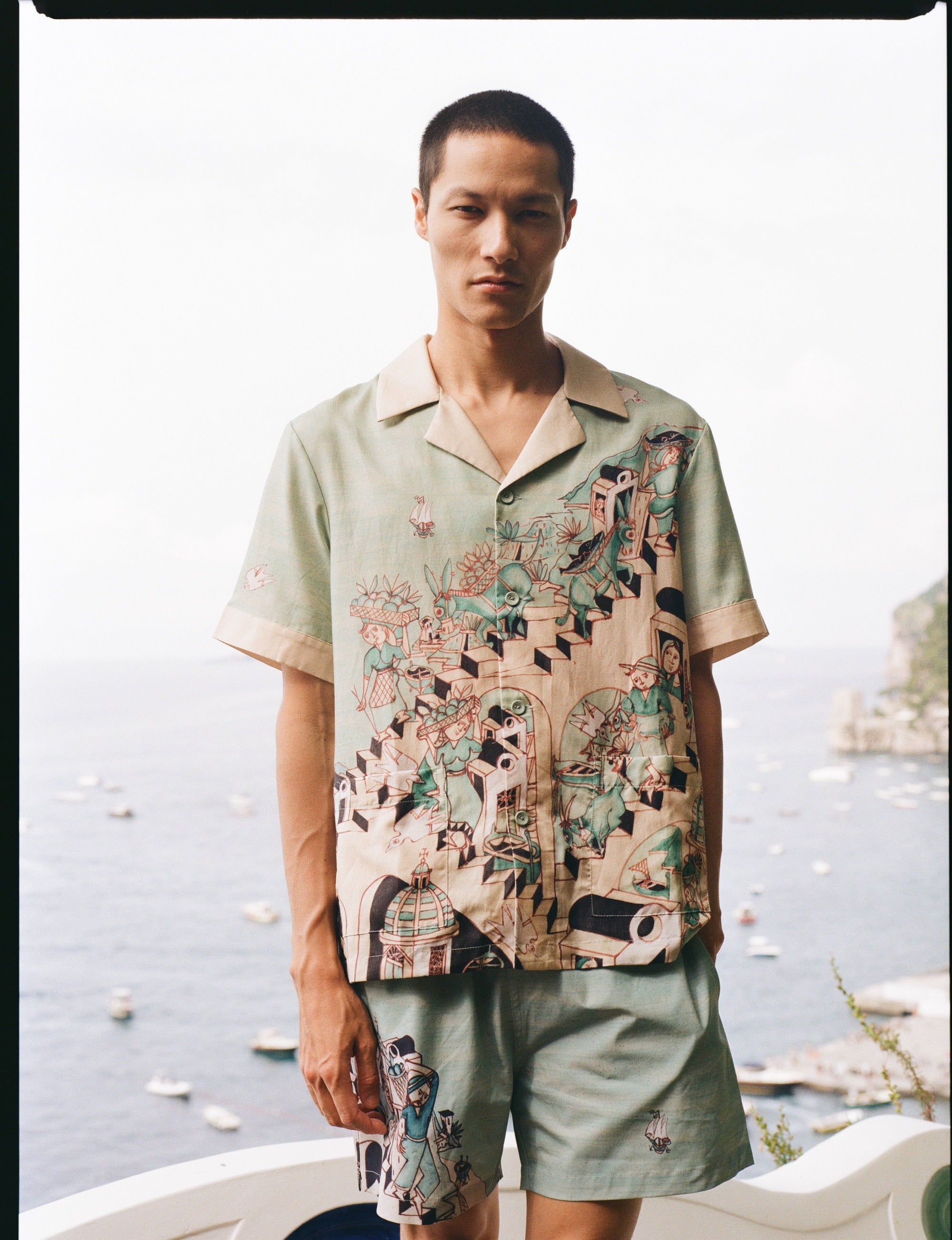 Man stares at camera with Amalfi bay in the background wearing the silk Liguori shirt and matching swim trunks
