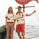 Woman and Man wearing the Da Adolfo shirts while standing on the dock next to the Da Adolfo restaurant sign, a red fish reflected in the embroidery