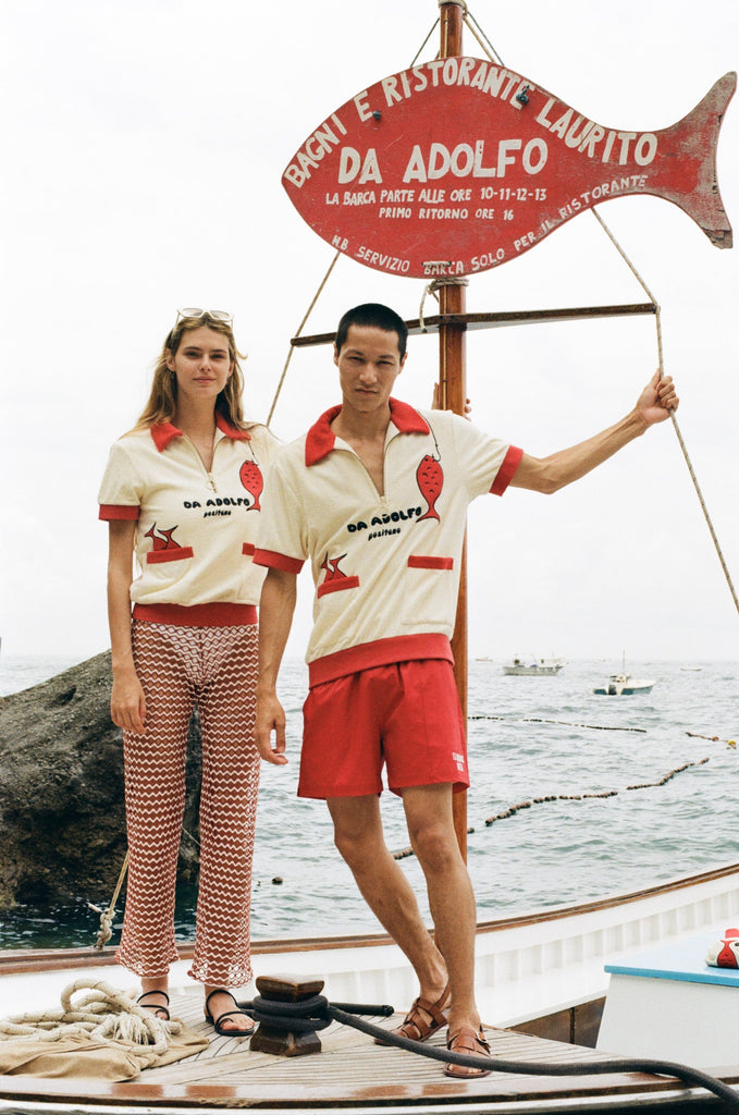 Woman and Man wearing the Da Adolfo shirts while standing on the dock next to the Da Adolfo restaurant sign, a red fish reflected in the embroidery