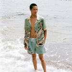 Man wearing unbuttoned Liguori shirt and matching swim trunks stands ankle deep in the ocean