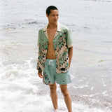 Man wearing unbuttoned Liguori shirt and matching swim trunks stands ankle deep in the ocean