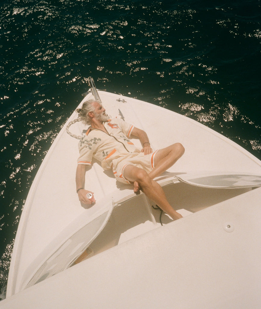 Man lounging on the bow of a white boat under the sun, wearing the matching 'Crouching Tiger' Cabana Set. He reclines with one arm behind his head and the other holding a can, surrounded by shimmering dark  ocean water.