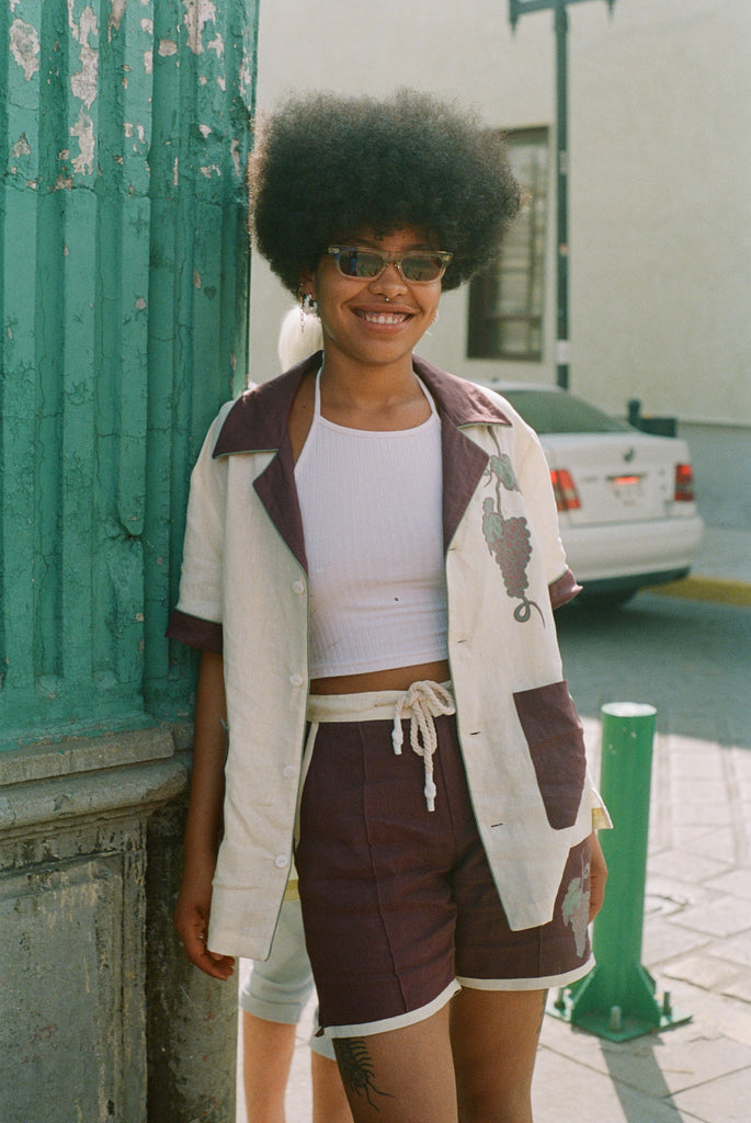Female model leaning against wall wearing full 'Tombarolo' cabana set. Shirt is open over a white tank top.