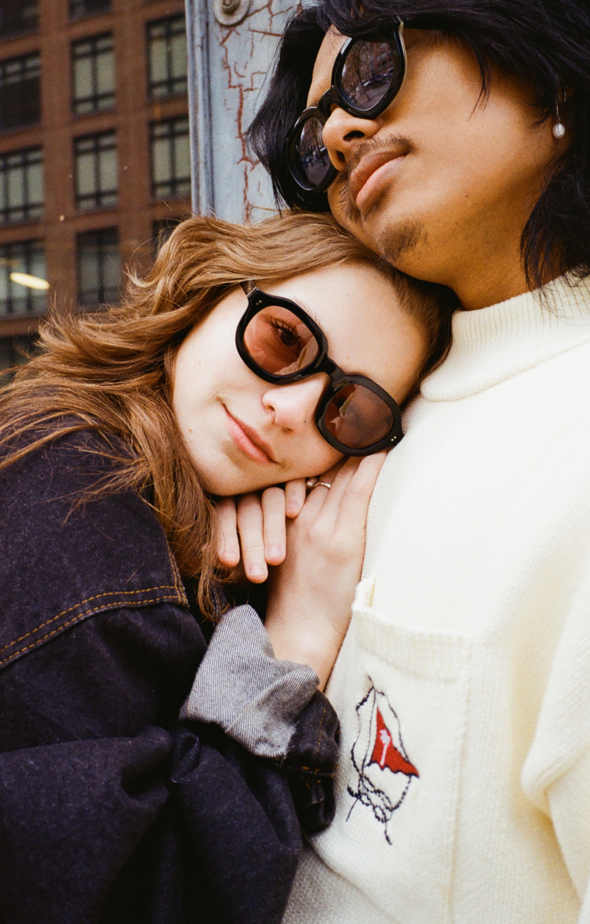 Young female model with reddish brown hair  leaning against model with black shoulder-length hair while the female model is wearing Lesca Lunetier x Tombolo lenses in coral and a denim jacket while the male model wears Lesca Lunetier X Tombolo lenses in charcoal and a white turtleneck with a background of a high-rise building's windows.