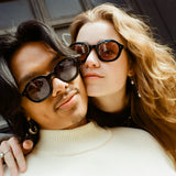 Male with black shoulder-length hair and Female model with reddish-brown hair standing cheek to cheek with the male wearing Lesca Lunetier x Tombolo Charcoal lenses and a white turtleneck and the female wearing Lesca Lunetier X Tombolo Coral lenses with a jean jacket.