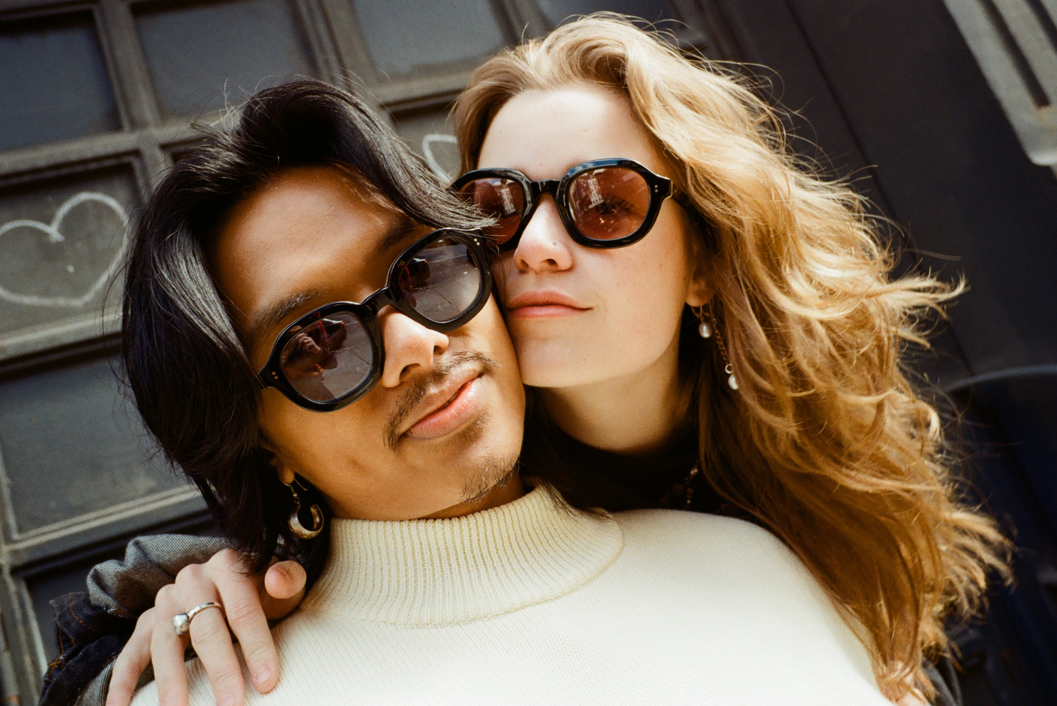 Male with black shoulder-length hair and Female model with reddish-brown hair standing cheek to cheek with the male wearing Lesca Lunetier x Tombolo Charcoal lenses and a white turtleneck and the female wearing Lesca Lunetier X Tombolo Coral lenses with a jean jacket.
