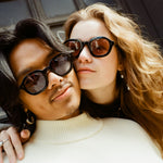 Male with black shoulder-length hair and Female model with reddish-brown hair standing cheek to cheek with the male wearing Lesca Lunetier x Tombolo Charcoal lenses and a white turtleneck and the female wearing Lesca Lunetier X Tombolo Coral lenses with a jean jacket.