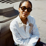 Young female model wearing Lesca Lunetier X Tombolo Glasses in Charcoal while seated wearing Sun Leapord Long-sleeve Cabana and holding a Coca-Cola in Washington Square Park.
