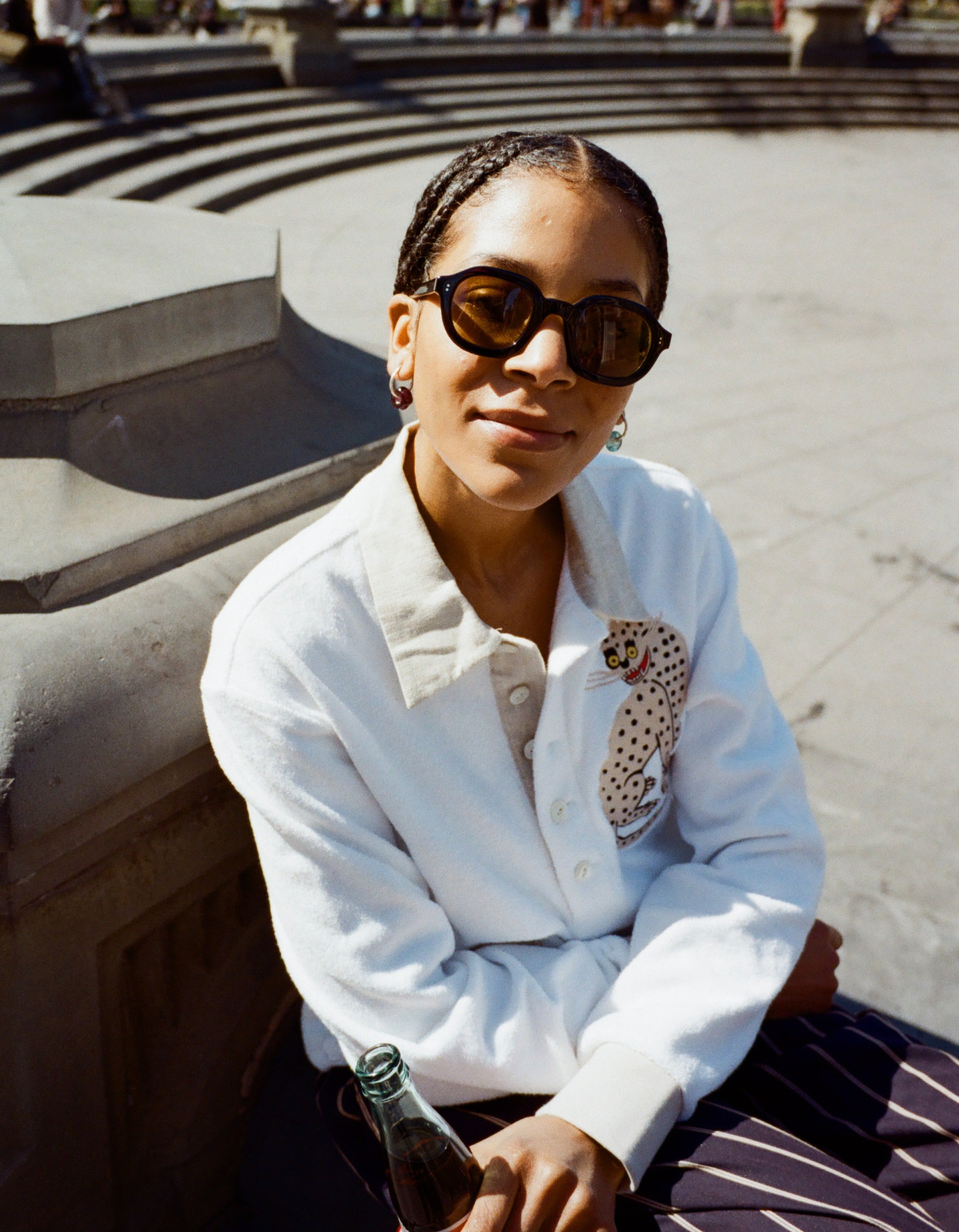 Young female model wearing Lesca Lunetier X Tombolo Glasses in Charcoal while seated wearing Sun Leapord Long-sleeve Cabana and holding a Coca-Cola in Washington Square Park.