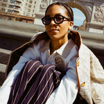 girl sitting in front of washington square park arch wearing black thick-framed sunglasses; also wearing beige jacket, white sweater, and navy striped pants