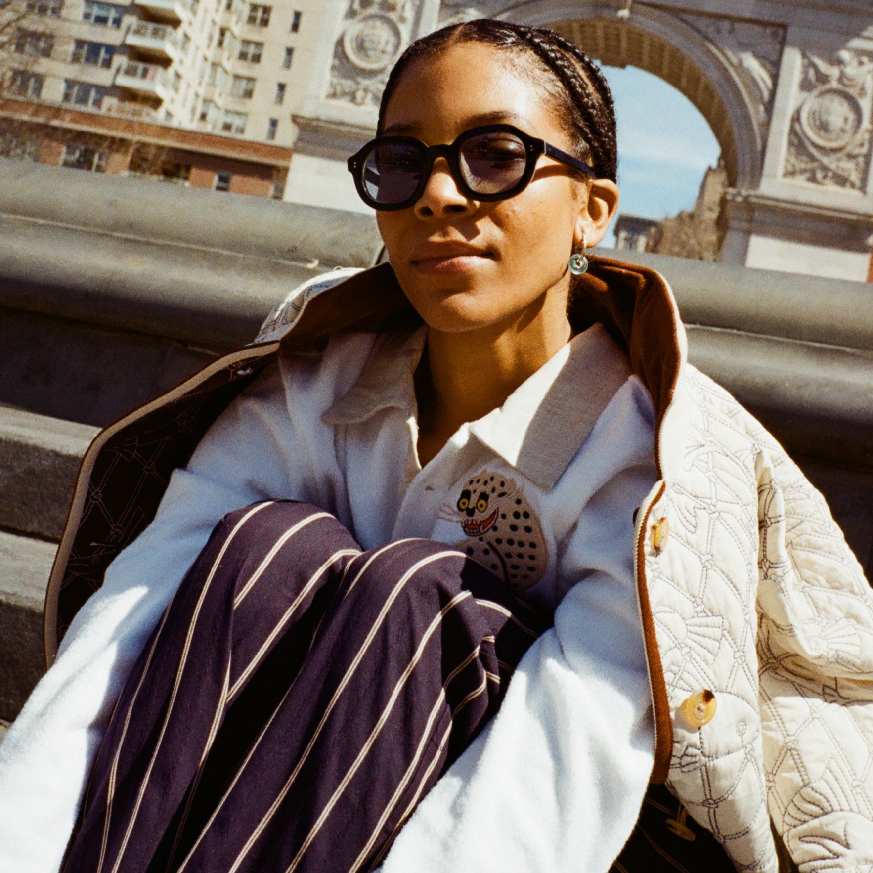 girl sitting in front of washington square park arch wearing black thick-framed sunglasses; also wearing beige jacket, white sweater, and navy striped pants