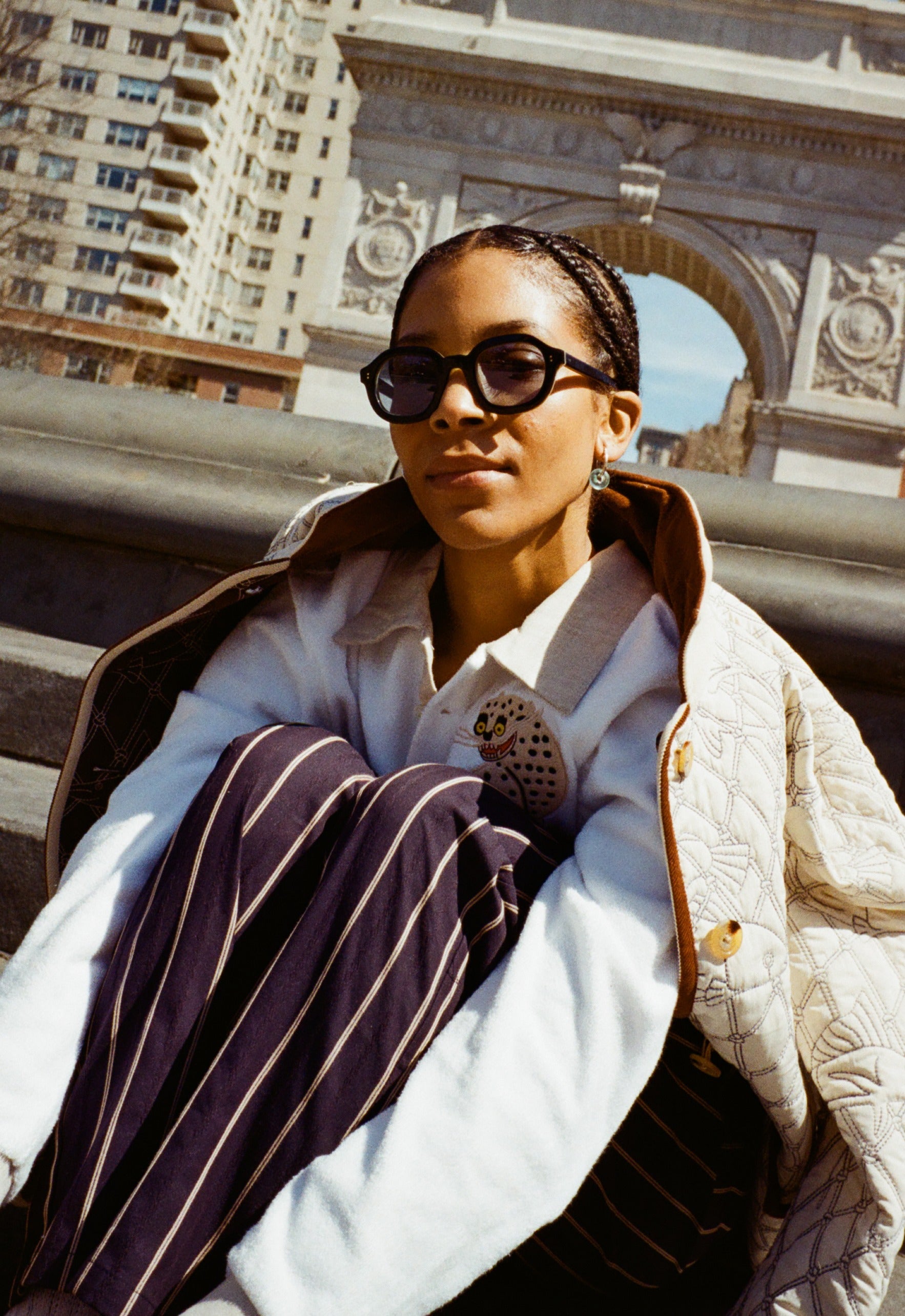 girl sitting in front of washington square park arch wearing black thick-framed sunglasses; also wearing beige jacket, white sweater, and navy striped pants