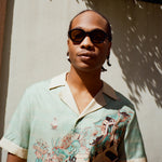 Young male model wearing Lesca Lunetier x Tombolo lenses in coral and a Tombolo X Le Sirenuse Hawaiian by Lucio Liguori on a beige background with trees appearing in the top.