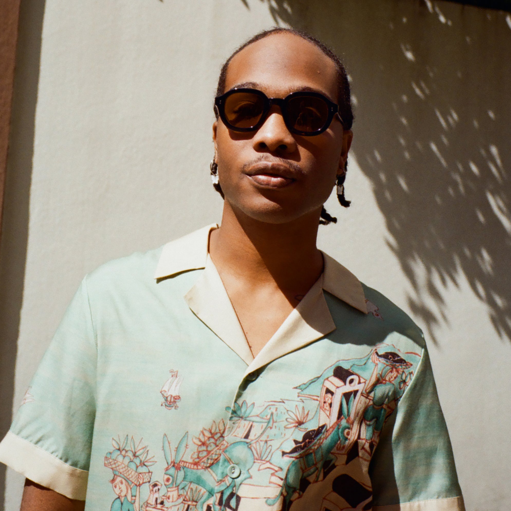 Young male model wearing Lesca Lunetier x Tombolo lenses in coral and a Tombolo X Le Sirenuse Hawaiian by Lucio Liguori on a beige background with trees appearing in the top.