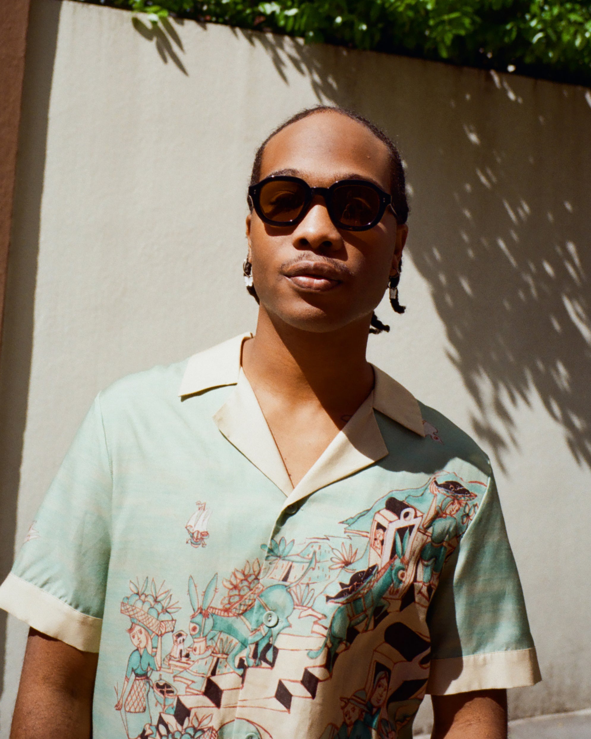 Young male model wearing Lesca Lunetier x Tombolo lenses in coral and a Tombolo X Le Sirenuse Hawaiian by Lucio Liguori on a beige background with trees appearing in the top.