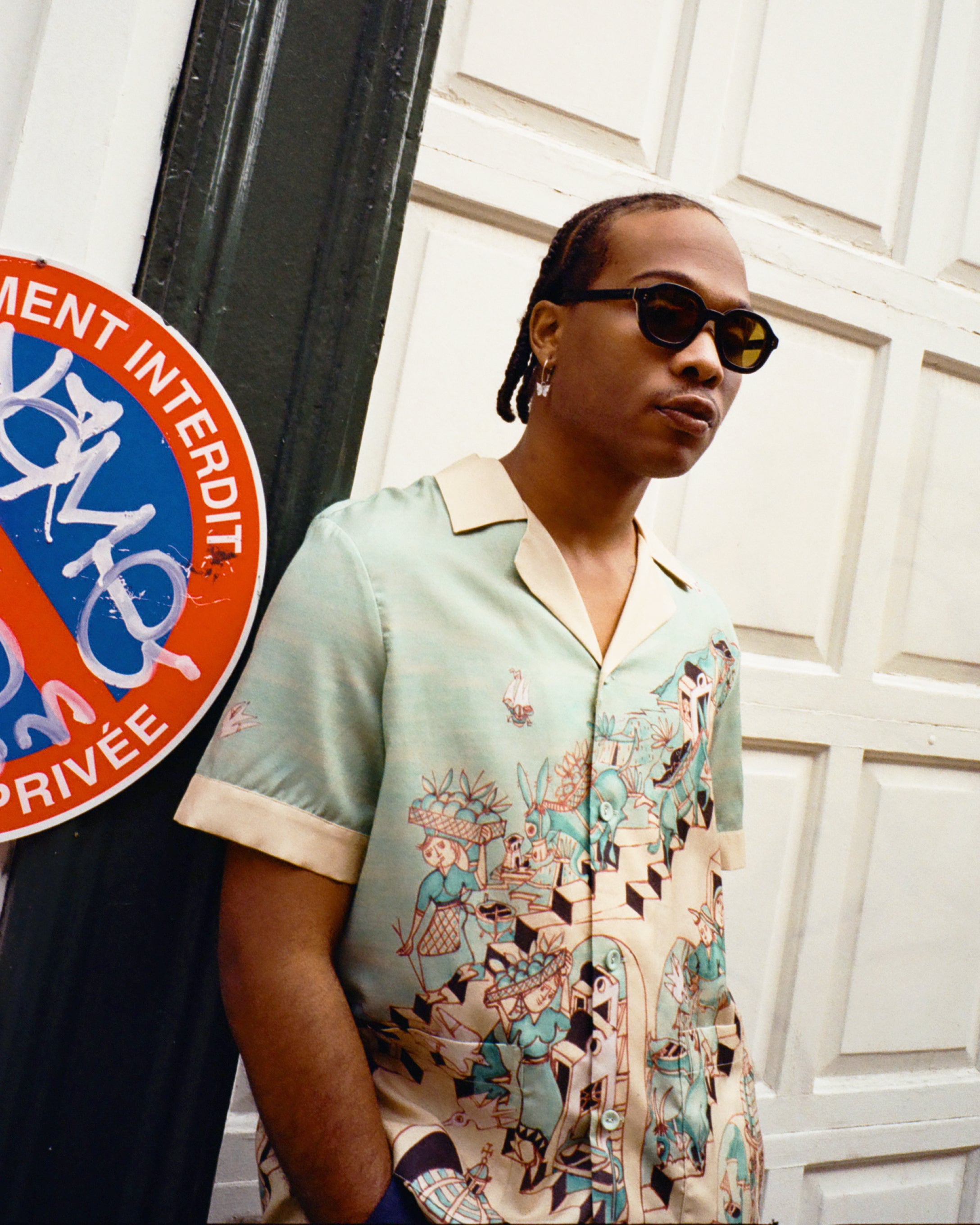 Young male model wearing Lesca Lunetier X Tombolo lenses in charcoal in addition to the "Le Sirenuse by Lucio Liguori" Hawaiian on the streets of Paris standing next to a No Parking sign written in french.