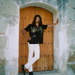Man standing in a stone doorway with one arm outstretched and the other on his hip. He wears the 'Big Brut' Cabana paired with white pants and black loafers. The background features a rustic wooden door framed by weathered stone walls.