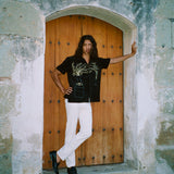 Man standing in a stone doorway with one arm outstretched and the other on his hip. He wears the 'Big Brut' Cabana paired with white pants and black loafers. The background features a rustic wooden door framed by weathered stone walls.