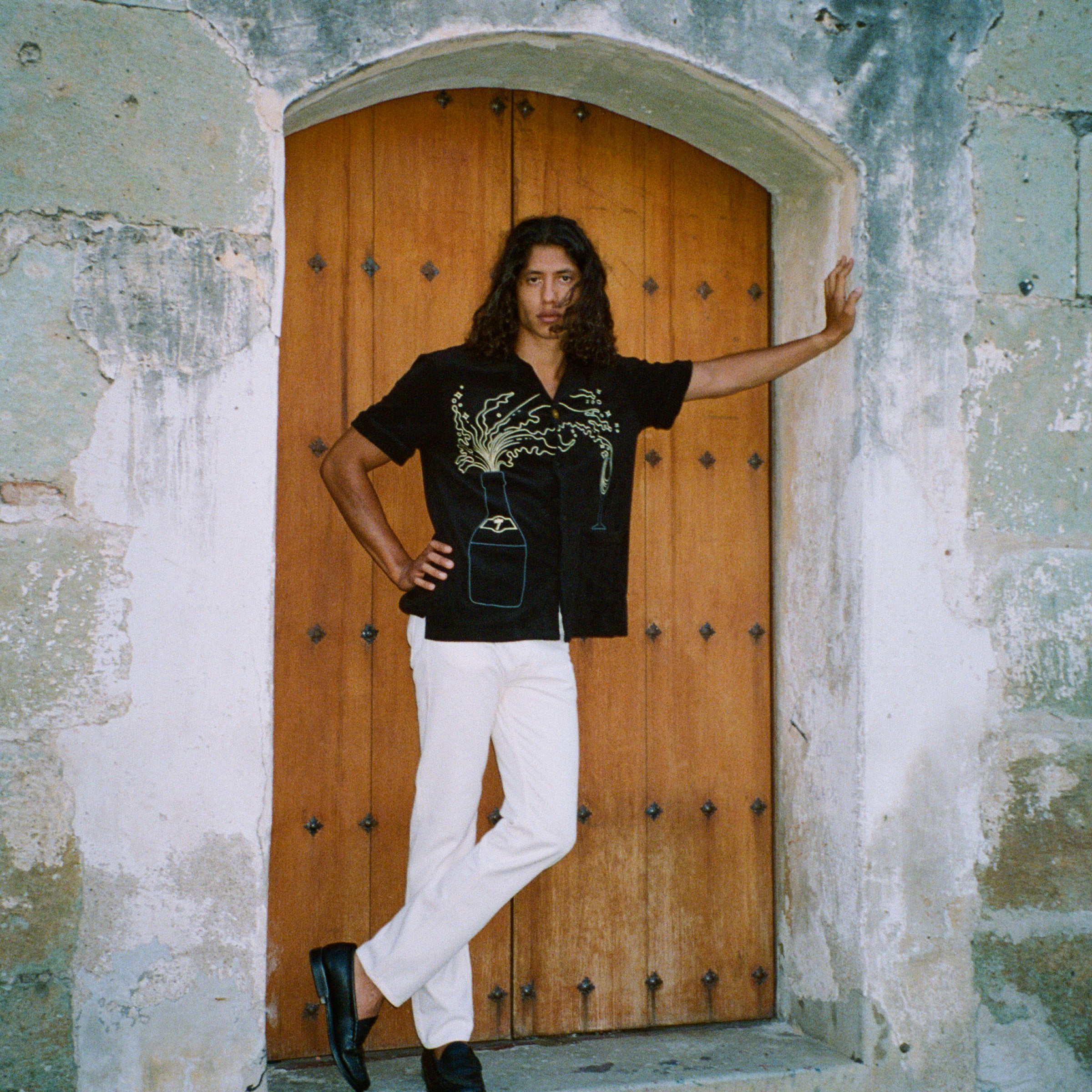Man standing in a stone doorway with one arm outstretched and the other on his hip. He wears the 'Big Brut' Cabana paired with white pants and black loafers. The background features a rustic wooden door framed by weathered stone walls.