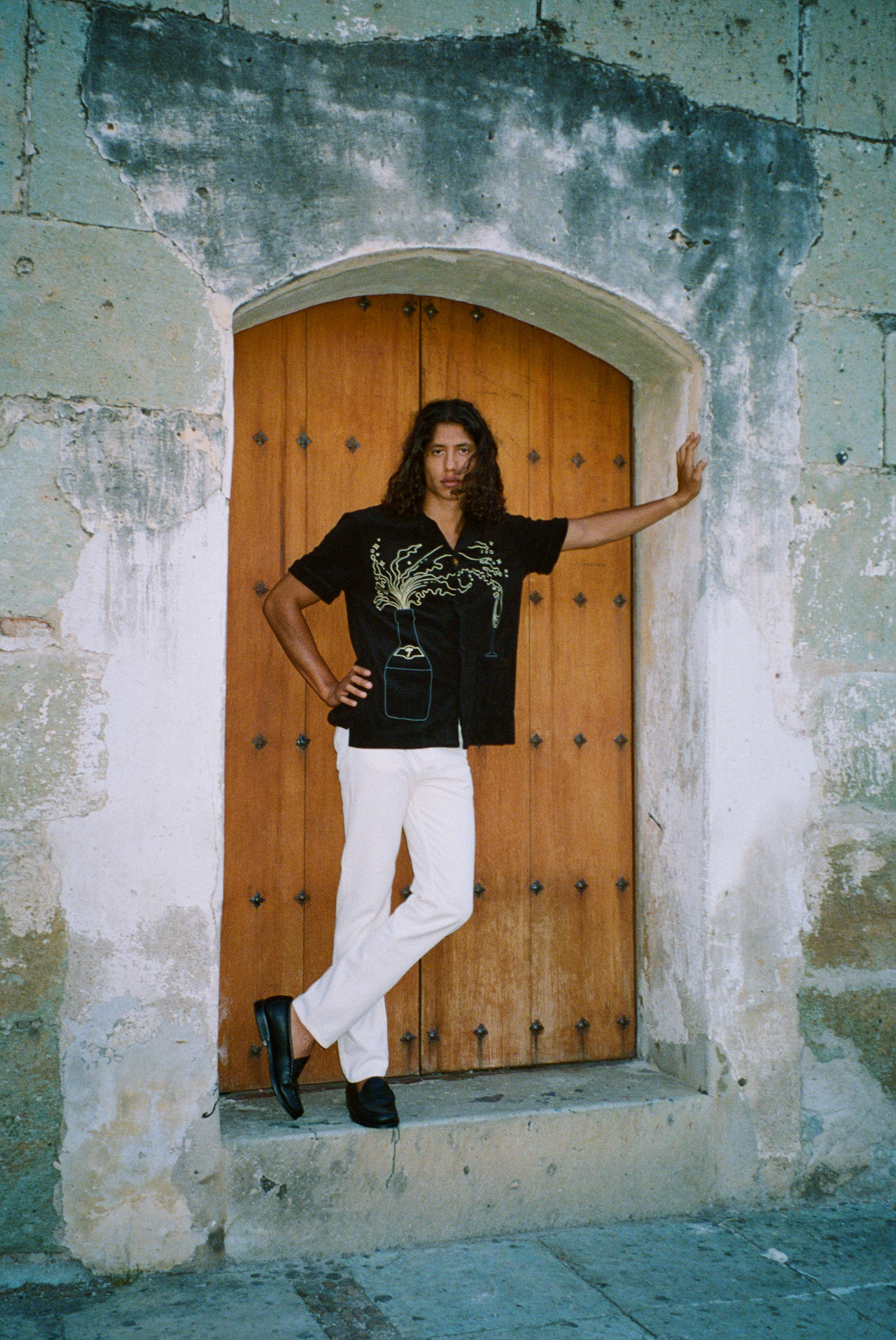 Man standing in a stone doorway with one arm outstretched and the other on his hip. He wears the 'Big Brut' Cabana paired with white pants and black loafers. The background features a rustic wooden door framed by weathered stone walls.