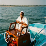 Woman on a light blue boat wearing a white hooded beach towel top with red trim and vertical 'Whaler' text. She stands behind a wooden console with a white steering wheel and retro controls.