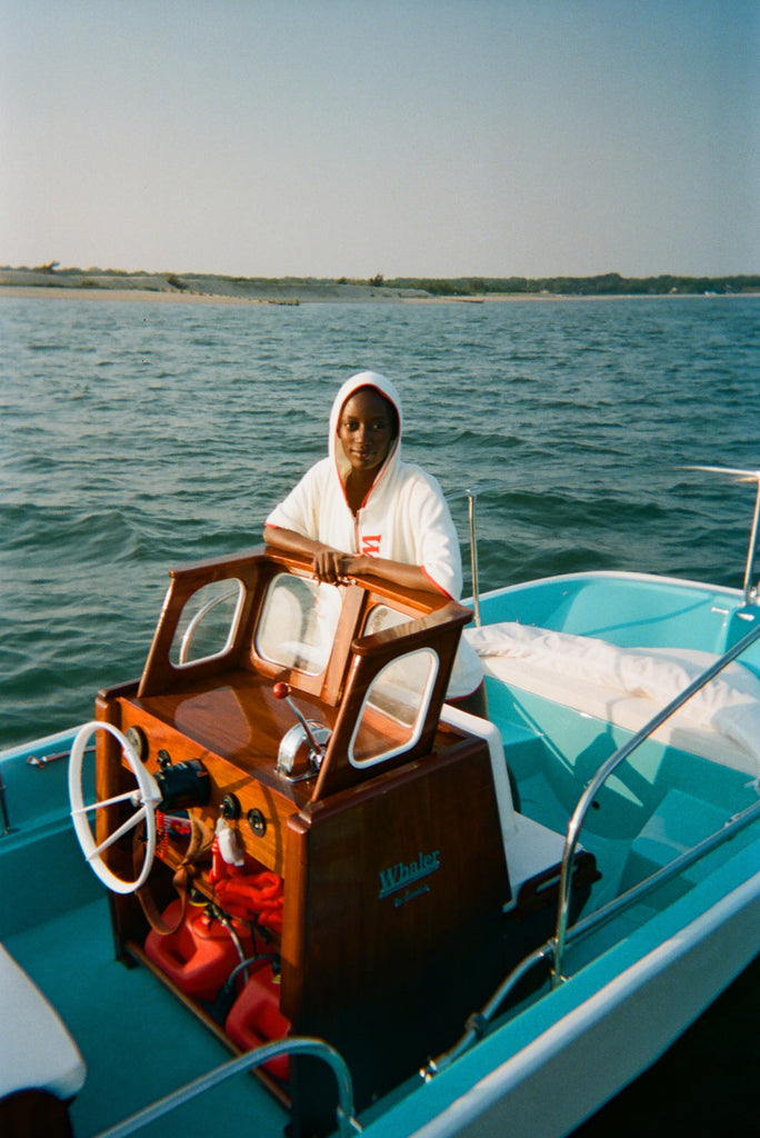 Woman on a light blue boat wearing a white hooded beach towel top with red trim and vertical 'Whaler' text. She stands behind a wooden console with a white steering wheel and retro controls.