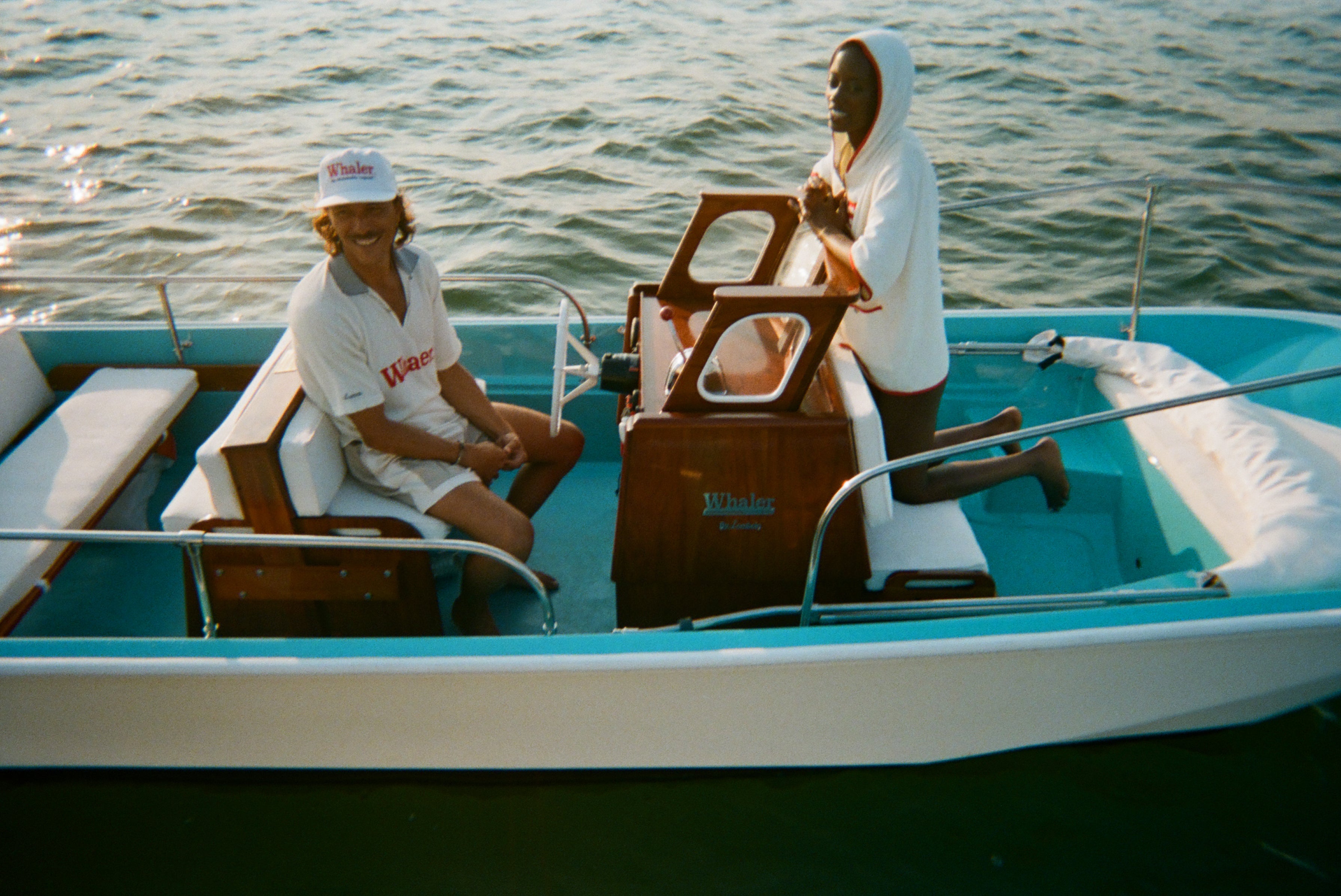 Two people sitting on a light blue and white boat. On the left, a man wears a white 'Whaler' polo shirt, white 'Whaler' cap, and shorts, smiling while seated at the steering bench. On the right, a woman kneels on a bench wearing a white terry zip-up with red trim and vertical 'Whaler' text on the front. 