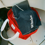 Navy board shorts with white contrast stitching and white 'Whaler' text on the leg, resting on top of a red canvas tote bag.