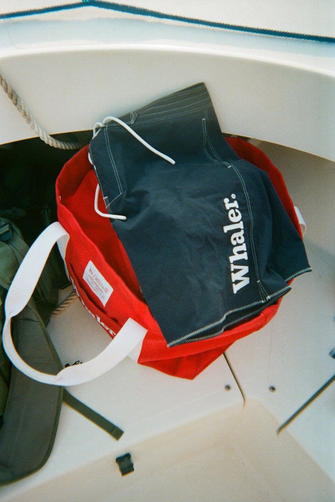 Navy board shorts with white contrast stitching and white 'Whaler' text on the leg, resting on top of a red canvas tote bag.