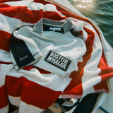 Light blue cotton mesh shirt with navy shoulder panels, a striped collar, and a 'Boston Whaler' logo on the chest, folded on top of a red and white striped towel on a boat. 