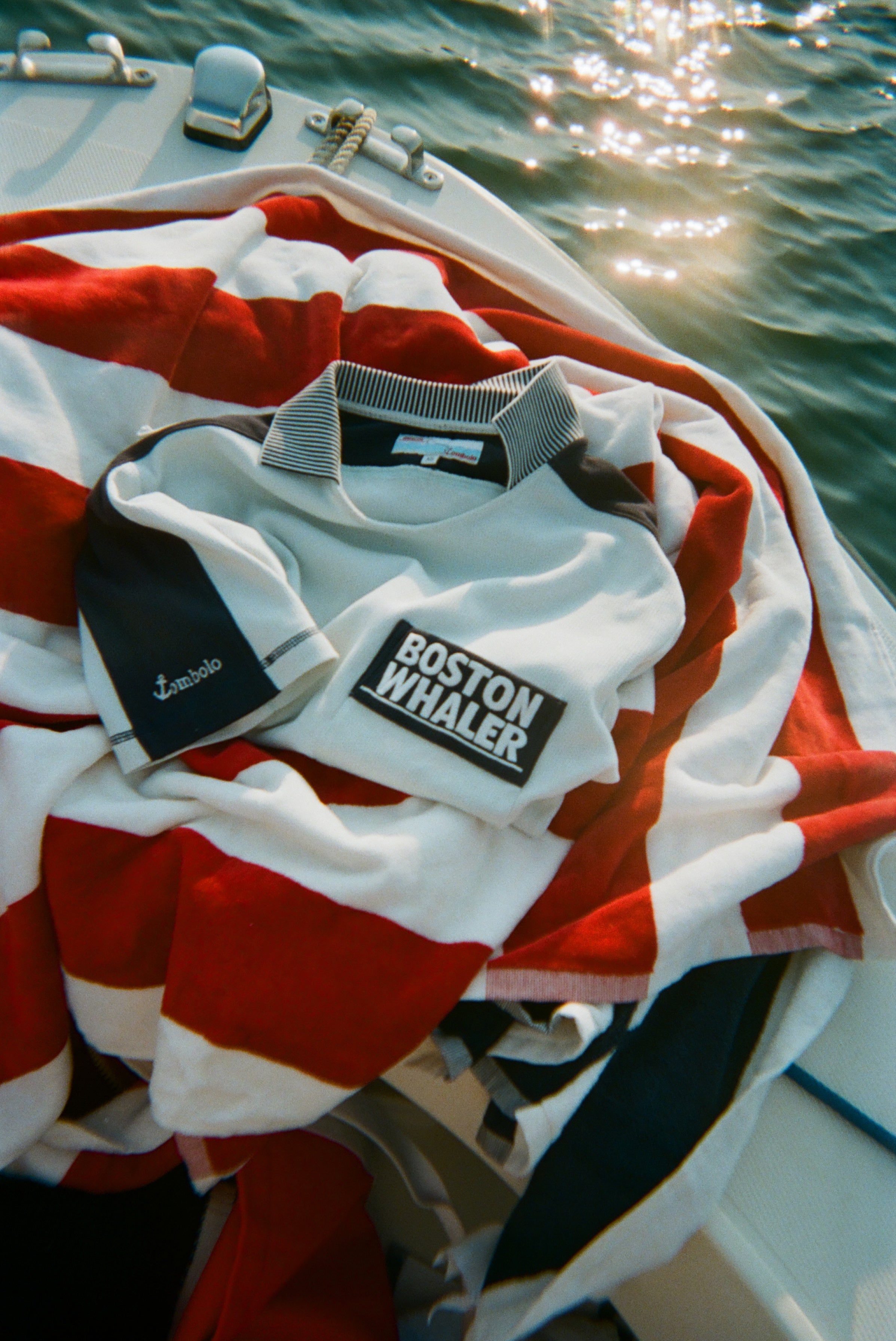 Light blue cotton mesh shirt with navy shoulder panels, a striped collar, and a 'Boston Whaler' logo on the chest, folded on top of a red and white striped towel on a boat. 