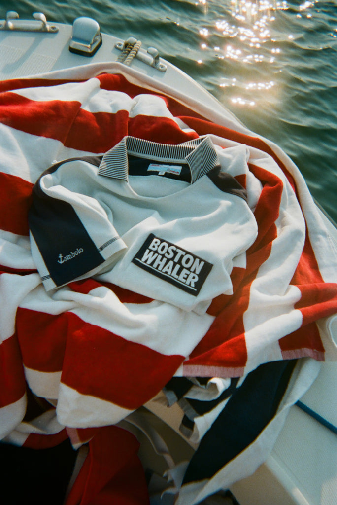 Light blue cotton mesh shirt with navy shoulder panels, a striped collar, and a 'Boston Whaler' logo on the chest, folded on top of a red and white striped towel on a boat. 
