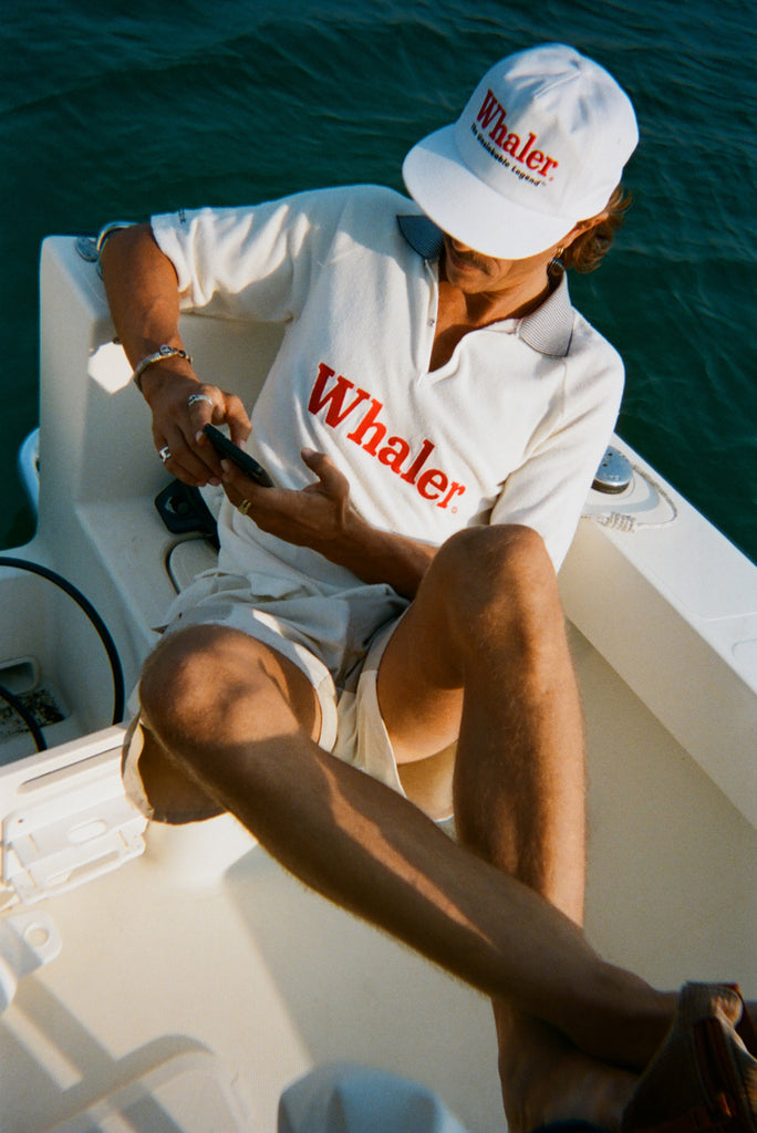 Man sitting on a boat while looking at his phone, wearing a white five-panel cap with red embroidered text reading 'Whaler' and smaller text reading 'The Unsinkable Legend'. He also wears a white collared shirt with red 'Whaler' text across the chest and cream-colored shorts.