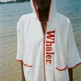 Woman standing in the ocean wearing a white hooded beach towel top with red trim, front pockets, and vertical red 'Whaler' text along the zipper.