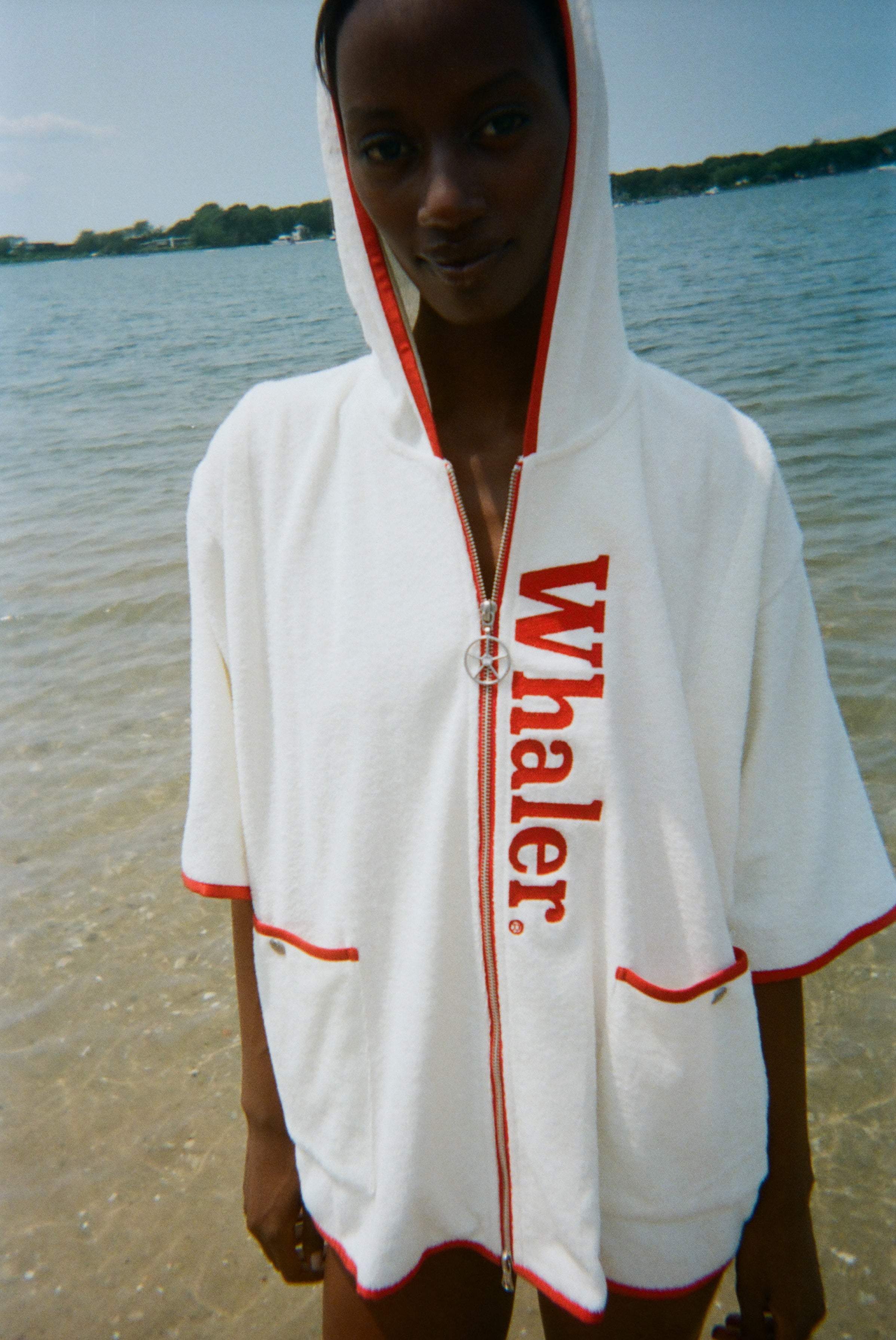Woman standing in the ocean wearing a white hooded beach towel top with red trim, front pockets, and vertical red 'Whaler' text along the zipper.