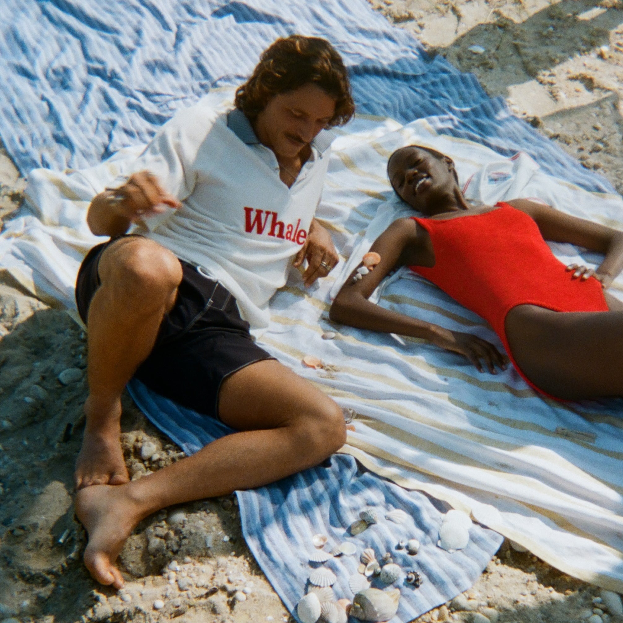 Two people laying out on towels at the beach. The man is wearing the white terry polo with 'Whaler' across the chest, and dark swim trunks. The woman is lying next to him in a red one-piece swimsuit. 