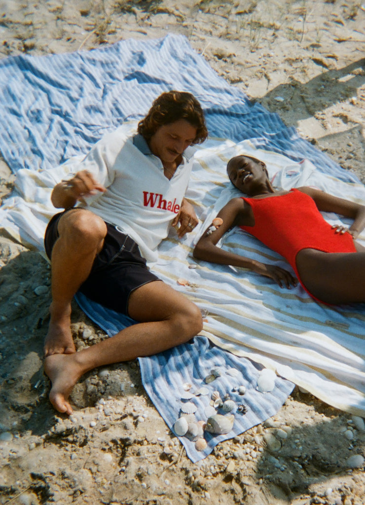 Two people laying out on towels at the beach. The man is wearing the white terry polo with 'Whaler' across the chest, and dark swim trunks. The woman is lying next to him in a red one-piece swimsuit. 