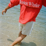 Man walking barefoot in shallow water, wearing two-tone khaki and off-white shorts and a red shirt with large white 'Boston Whaler' text across the back flap.