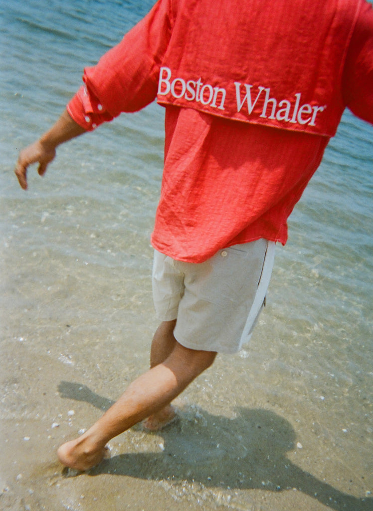 Man walking barefoot in shallow water, wearing two-tone khaki and off-white shorts and a red shirt with large white 'Boston Whaler' text across the back flap.