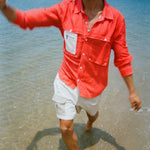 A man splashes through shallow water wearing a bright red button-up shirt with a white mesh zip pocket layered on the left chest. He pairs it with khaki and off white pleated shorts featuring a small 'Whaler' logo patch on the pocket.