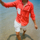 A man splashes through shallow water wearing a bright red button-up shirt with a white mesh zip pocket layered on the left chest. He pairs it with khaki and off white pleated shorts featuring a small 'Whaler' logo patch on the pocket.