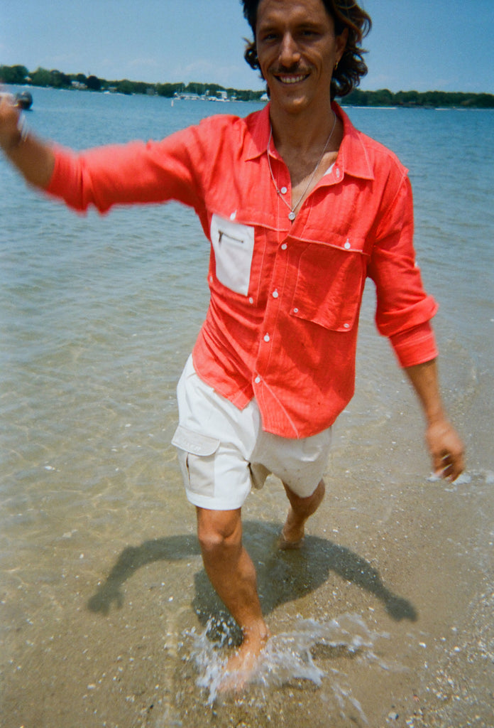 A man splashes through shallow water wearing a bright red button-up shirt with a white mesh zip pocket layered on the left chest. He pairs it with khaki and off white pleated shorts featuring a small 'Whaler' logo patch on the pocket.