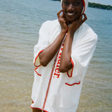Woman standing in the ocean, smiling,  wearing a white hooded beach towel top with red trim, front pockets, and vertical red 'Whaler' text along the zipper.