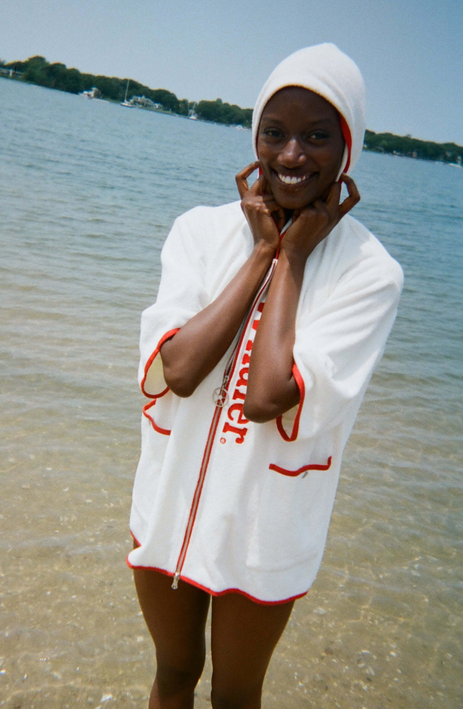 Woman standing in the ocean, smiling,  wearing a white hooded beach towel top with red trim, front pockets, and vertical red 'Whaler' text along the zipper.