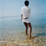 Woman standing in shallow water with her back to us, wearing the white terry short-sleeve 'Boston Whaler' Cabana, which features a boat illustration on the back.