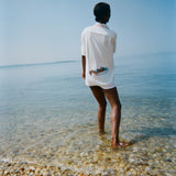 Woman standing in shallow water with her back to us, wearing the white terry short-sleeve 'Boston Whaler' Cabana, which features a boat illustration on the back.